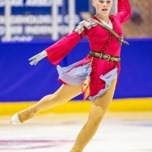 May include: A young figure skater wearing a red and blue costume with gold accents performs a move on the ice. The costume features a long, flowing skirt and a belt with a buckle.