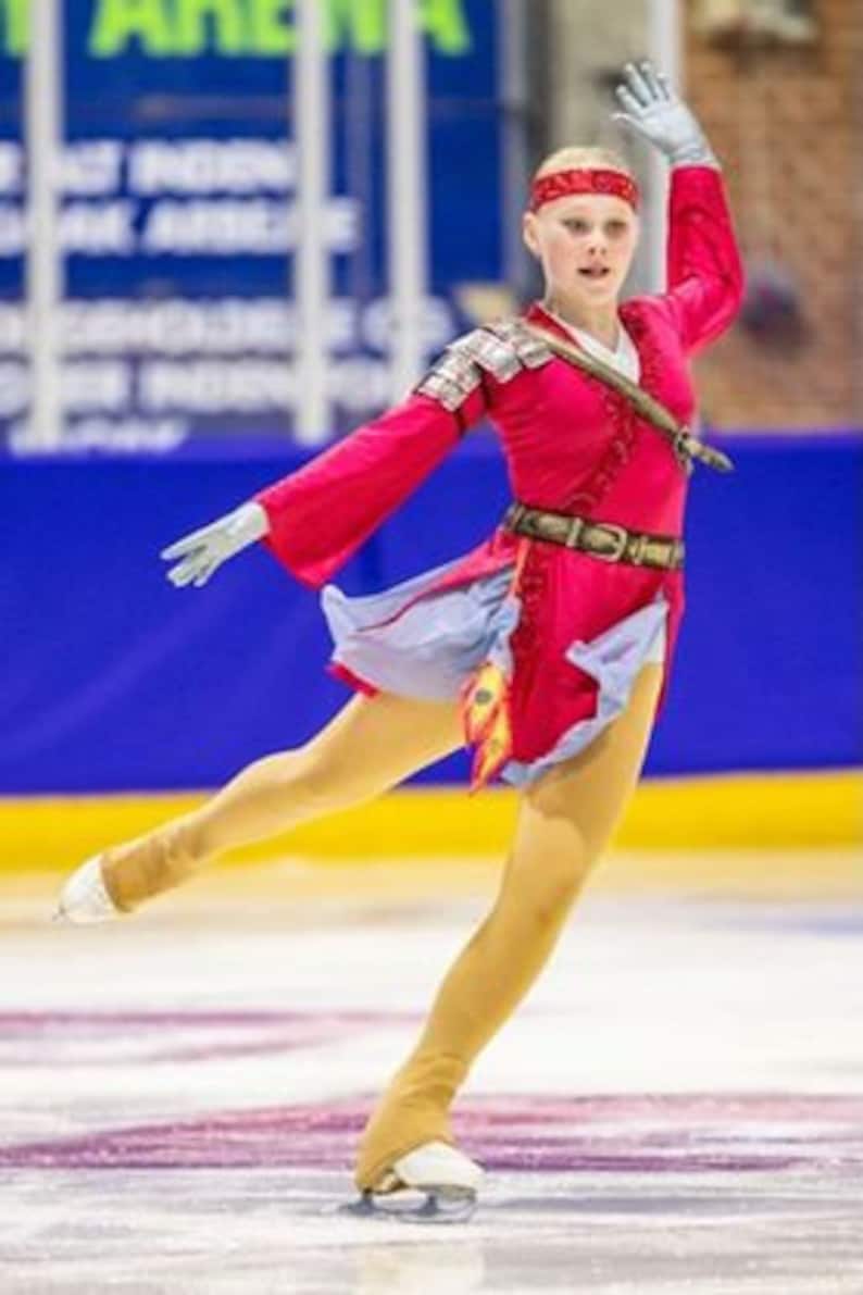 May include: A young figure skater wearing a red and blue costume with gold accents performs a move on the ice. The costume features a long, flowing skirt and a belt with a buckle.