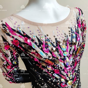 May include: A black and pink leotard with a geometric design featuring rhinestones and crystals. The leotard has long sleeves and a scoop neck.