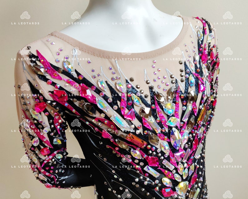 May include: A black and pink leotard with a geometric design featuring rhinestones and crystals. The leotard has long sleeves and a scoop neck.