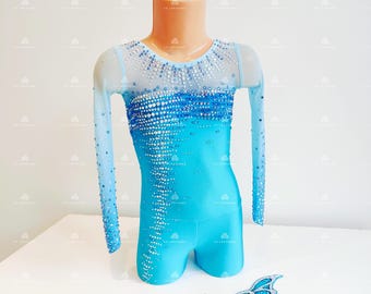Puede incluir: Maillot turquesa y azul con mangas largas, adornado con pedrería. El maillot se exhibe en un maniquí. Una pieza decorativa a juego se coloca debajo. La prenda está diseñada para gimnasia o danza.