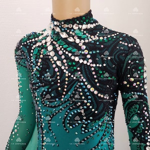 May include: A teal and black leotard with long sleeves, embellished with rhinestones and crystals. The bodice features a black lace overlay with a swirling pattern. The high neckline is also adorned with crystals.