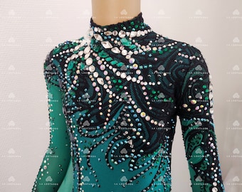 Puede incluir: Maillot de color verde azulado y negro con mangas largas, adornado con pedrería y cristales. El corpiño presenta una superposición de encaje negro con un diseño en espiral. El cuello alto también está adornado con cristales.