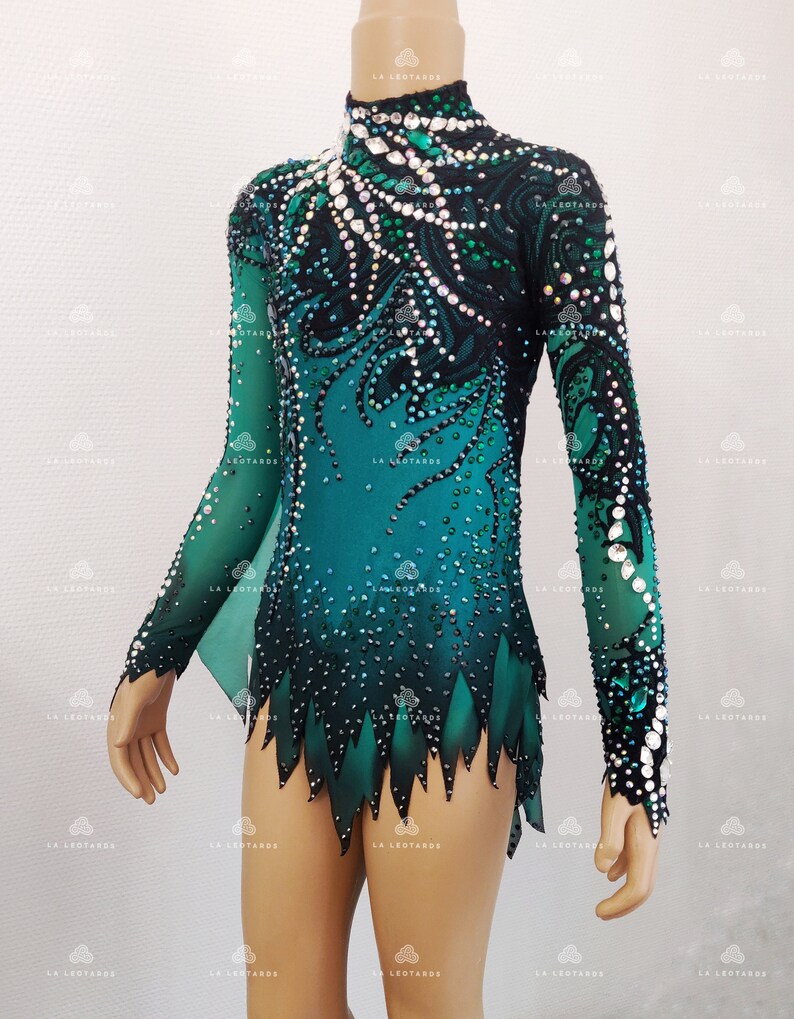 May include: A teal and black leotard with long sleeves, decorated with rhinestones. The design includes a high neckline and a decorative pattern flowing down the bodice. The sleeves are sheer, and the hem has a jagged edge. The brand name "LA LEOTARDS" is visible.