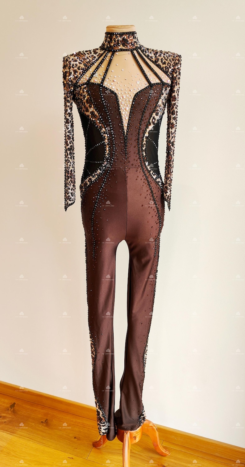 May include: A brown and black dance leotard with a leopard print design and rhinestones. The leotard has long sleeves and a high neckline.