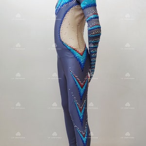 Op de afbeelding: Een grijs pakje met blauwe en turquoise accenten en strass. Het pakje heeft lange mouwen en een hoge hals. Het pakje is ontworpen voor kunstschaatsen.