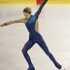 Op de afbeelding: Een vrouwelijke kunstschaatsster die een blauw en beige kunstschaatskostuum draagt met een blauw en wit ontwerp. Ze voert een beweging uit op het ijs met haar rechterbeen gestrekt en haar linkerbeen op een schaats.