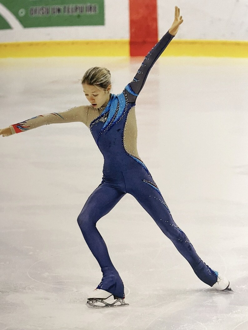 Op de afbeelding: Een vrouwelijke kunstschaatsster die een blauw en beige kunstschaatskostuum draagt met een blauw en wit ontwerp. Ze voert een beweging uit op het ijs met haar rechterbeen gestrekt en haar linkerbeen op een schaats.