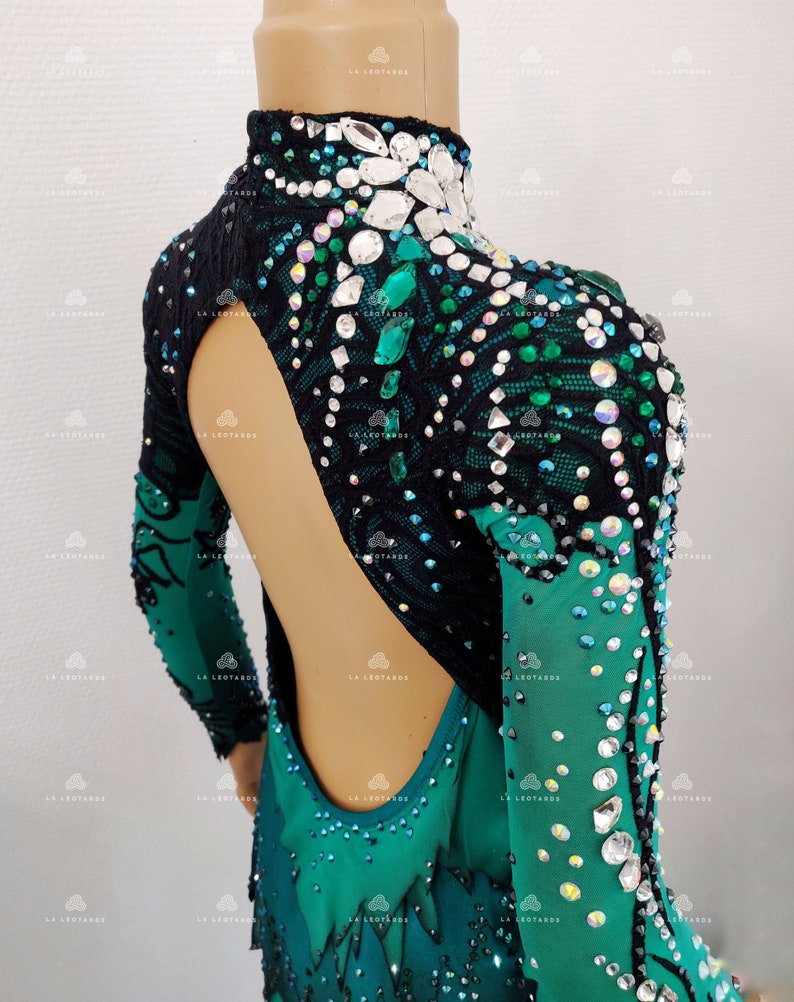 May include: A green and black leotard with long sleeves, adorned with rhinestones and crystals. The leotard has a high neck and an open back design. The sleeves have a gradient effect, changing from green to black. The garment is designed for dance or gymnastics.