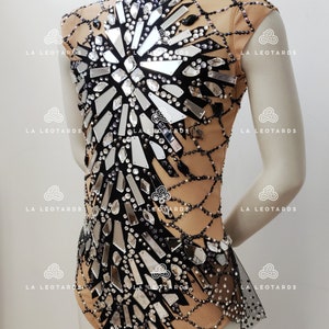 May include: A nude leotard with a black mesh overlay and a design of mirrored shards and rhinestones. The leotard has a high neck and short sleeves.