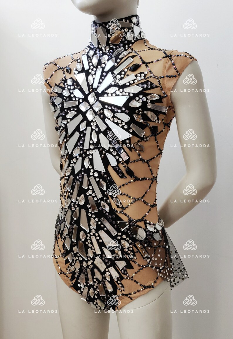 May include: A nude leotard with a black mesh overlay and a design of mirrored shards and rhinestones. The leotard has a high neck and short sleeves.