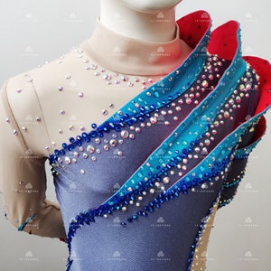 Op de afbeelding: Een blauwe, grijze en rode maillot met een hoge halslijn en lange mouwen. De maillot is versierd met strass en heeft een gerimpeld ontwerp op de schouder.