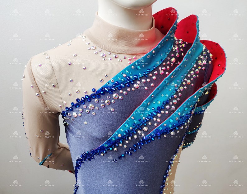 Op de afbeelding: Een blauwe, grijze en rode maillot met een hoge halslijn en lange mouwen. De maillot is versierd met strass en heeft een gerimpeld ontwerp op de schouder.