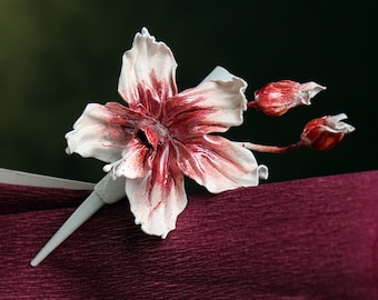 May include: A handcrafted floral brooch featuring a white and burgundy flower with detailed petals and two buds. The flower is mounted on a gray pin, resting on a textured, deep red surface. The brooch is a unique accessory.