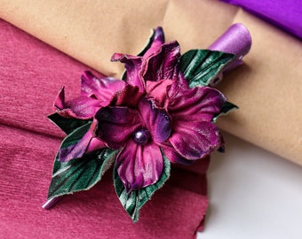 May include: A handcrafted, deep purple leather flower with green leaves and a central bead. The flower is set against a backdrop of textured maroon and brown paper, creating a striking contrast. The flower's petals have a gradient effect.