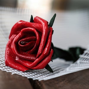 Peut inclure: Une rose rouge en cuir, avec une tige et des feuilles vertes, posée sur un treillis blanc. Les pétales de la rose sont détaillés et brillants. L'arrière-plan est flou, avec une surface en bois. Article fait à la main.