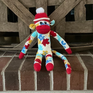 May include: A sock monkey wearing a red and white Santa hat with a white pom-pom. The monkey is sitting on a brick surface and has a white, blue, and yellow patterned body.