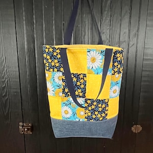 May include: A large tote bag with a patchwork design featuring yellow, blue, and white floral patterns. The bag has a denim-like bottom and navy blue handles and straps. The bag is hanging against a dark wooden background.