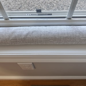 May include: A gray fabric draft stopper for a window. The draft stopper is placed under a white window sill. A white electrical outlet is visible below the sill.