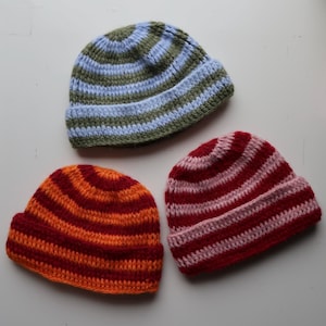 Könnte beinhalten: Drei gestreifte Strickmützen. Eine Mütze ist grün und blau, eine ist rot und orange und eine ist rosa und rot.