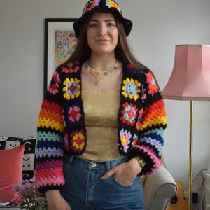 May include: A woman wearing a rainbow crochet top with a white crocheted bucket hat and blue jeans. The top features a black background with colorful squares.