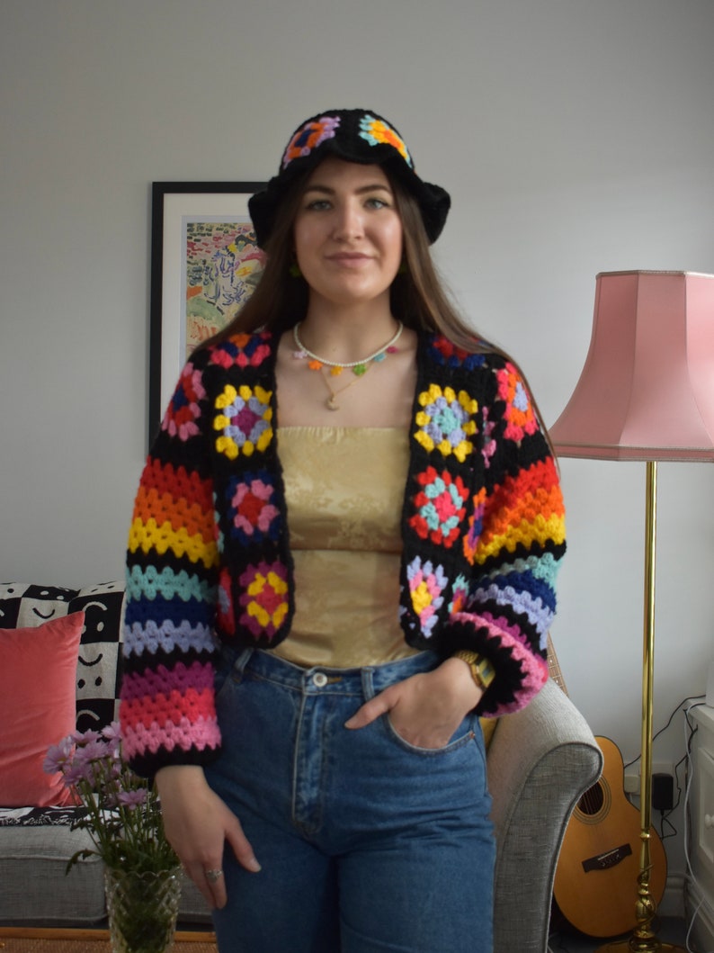 May include: A woman wearing a rainbow crochet top with a white crocheted bucket hat and blue jeans. The top features a black background with colorful squares.