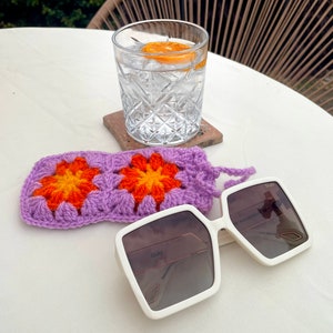 K&ouml;nnte beinhalten: Wei&szlig;e quadratische Sonnenbrille mit einem lila geh&auml;kelten Etui. Die Sonnenbrille hat eine braune T&ouml;nung und der Text "QUAY" ist auf dem Rahmen sichtbar. Das Etui hat ein Blumenmuster mit orangefarbenen und gelben Blumen.