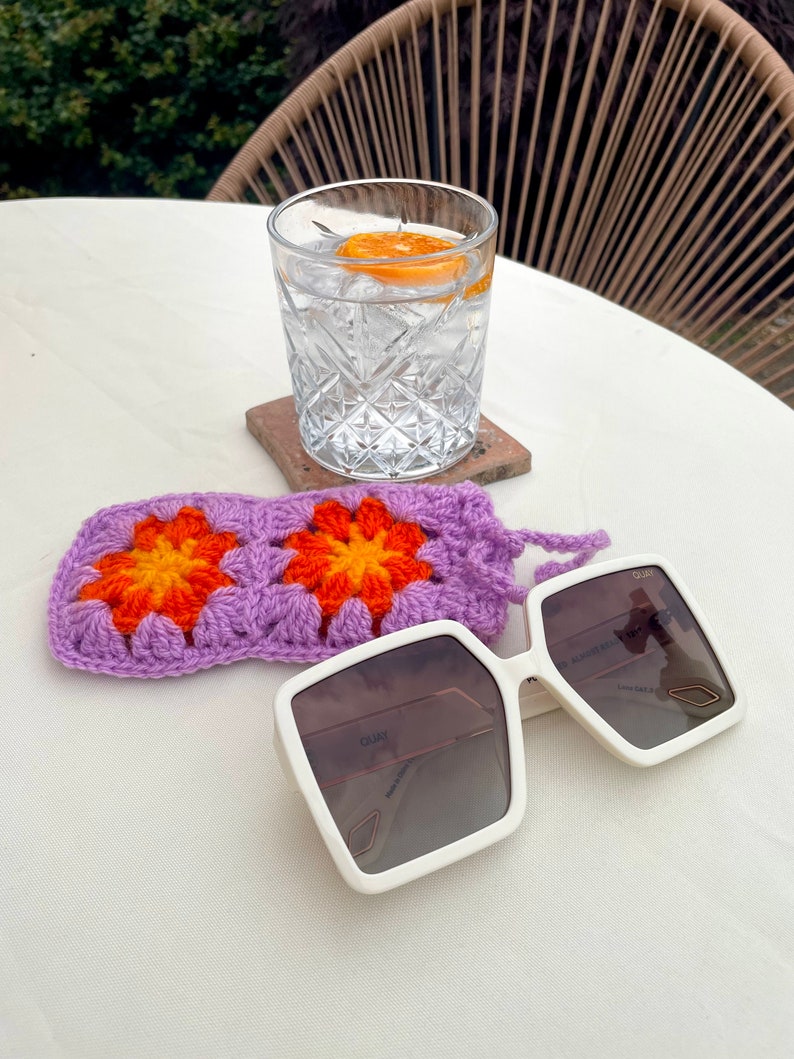 K&ouml;nnte beinhalten: Wei&szlig;e quadratische Sonnenbrille mit einem lila geh&auml;kelten Etui. Die Sonnenbrille hat eine braune T&ouml;nung und der Text "QUAY" ist auf dem Rahmen sichtbar. Das Etui hat ein Blumenmuster mit orangefarbenen und gelben Blumen.