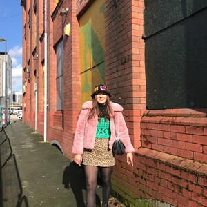 May include: A woman wearing a pink faux fur jacket, a green crochet top, a leopard print skirt, black tights, and black combat boots. She is also wearing a black bucket hat with colorful embroidery.