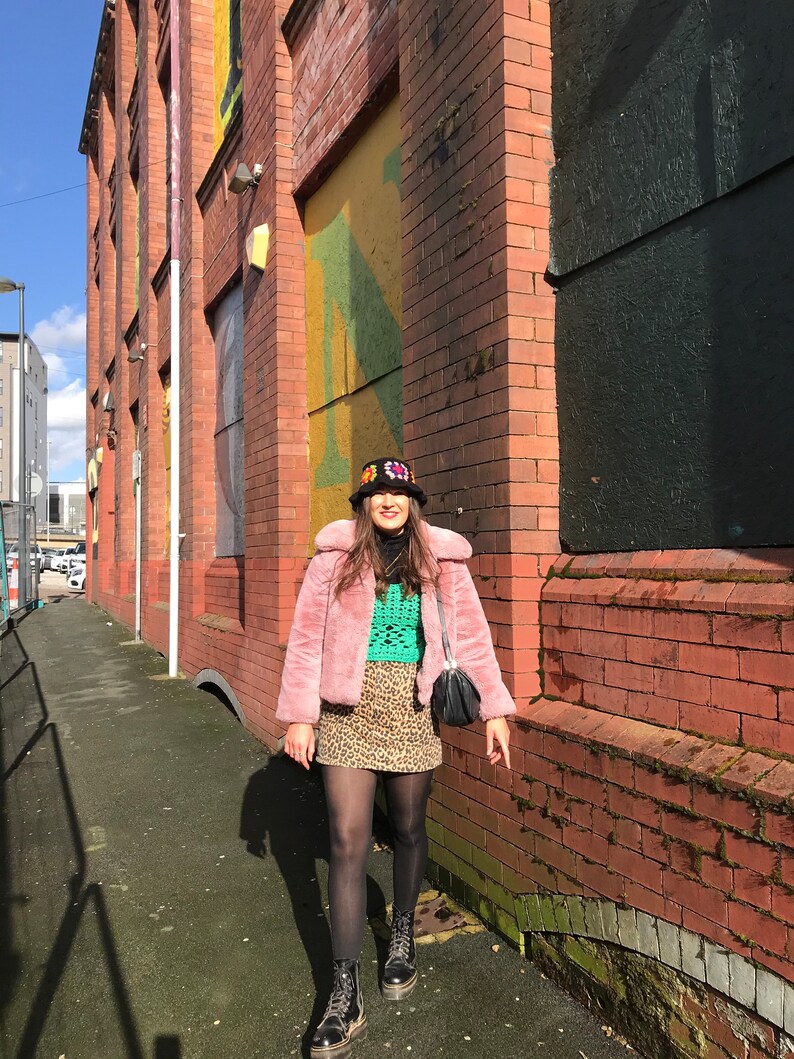 May include: A woman wearing a pink faux fur jacket, a green crochet top, a leopard print skirt, black tights, and black combat boots. She is also wearing a black bucket hat with colorful embroidery.