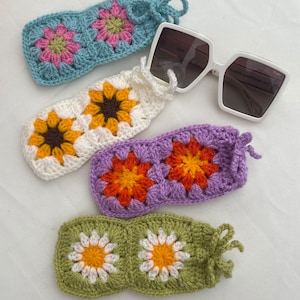 K&ouml;nnte beinhalten: Vier geh&auml;kelte Brillenetuis mit verschiedenen Blumendesigns. Die Etuis sind mit einem Granny-Square-Stich gearbeitet und in verschiedenen Farben gehalten, darunter Blau, Wei&szlig;, Lila und Gr&uuml;n. Jedes Etui hat einen kleinen Quasten am Ende.