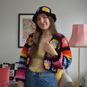 May include: A woman wearing a rainbow crochet top with a white crocheted hat and blue jeans.