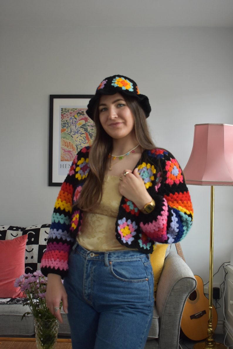May include: A woman wearing a rainbow crochet top with a white crocheted hat and blue jeans.