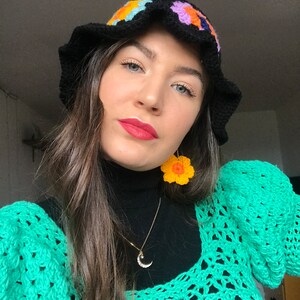 May include: A woman wearing a black crochet bucket hat with colorful flower accents, a black turtleneck sweater, a gold crescent moon necklace, and a green crochet sweater with large sleeves. She is also wearing yellow crochet flower earrings.