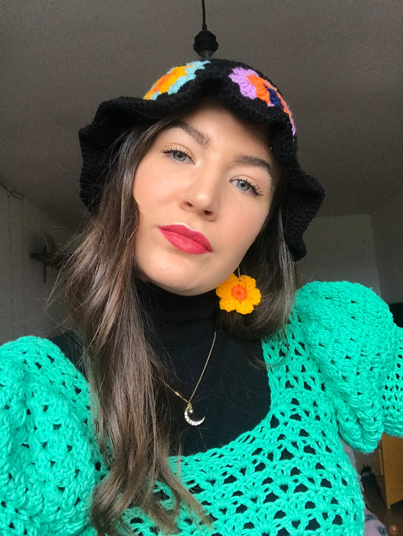 May include: A woman wearing a black crochet bucket hat with colorful flower accents, a black turtleneck sweater, a gold crescent moon necklace, and a green crochet sweater with large sleeves. She is also wearing yellow crochet flower earrings.