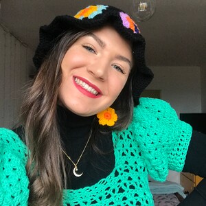 May include: A woman wearing a black crocheted bucket hat with colorful flower accents, a green crochet top, and a gold necklace with a crescent moon pendant. She is also wearing yellow flower-shaped earrings.