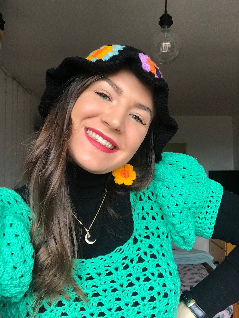 May include: A woman wearing a black crocheted bucket hat with colorful flower accents, a green crochet top, and a gold necklace with a crescent moon pendant. She is also wearing yellow flower-shaped earrings.