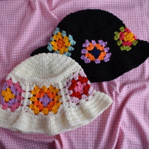 May include: Two crocheted bucket hats, one white with colorful granny squares and one black with purple, blue, and green granny squares.