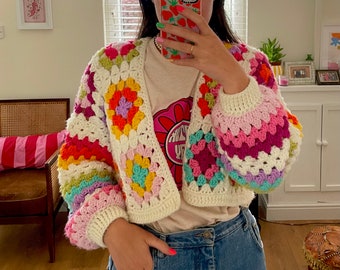 May include: A cropped, rainbow crochet cardigan with a white background and colorful granny squares. The cardigan is worn over a white t-shirt with a graphic print.