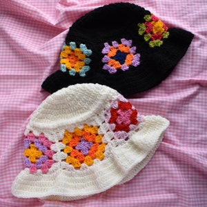 May include: Two crocheted bucket hats with colorful granny square designs. One hat is black with multi-colored squares, and the other is white with pink, orange, and purple squares.