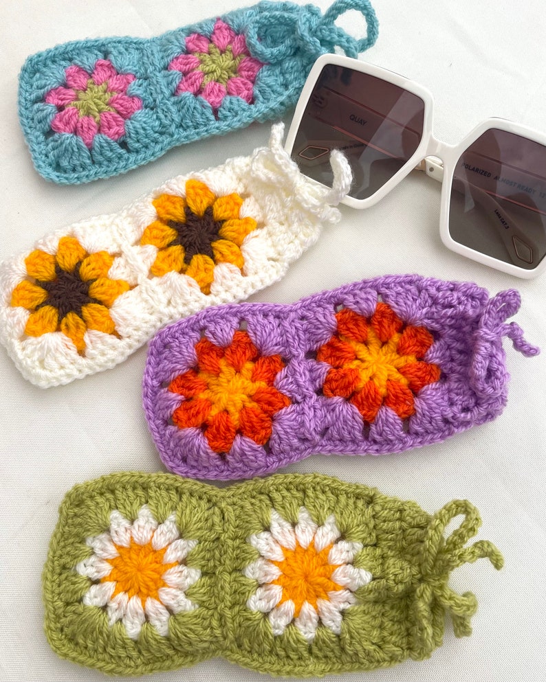 K&ouml;nnte beinhalten: Vier geh&auml;kelte Brillenetuis mit verschiedenen Blumendesigns. Die Etuis sind in Blau, Wei&szlig;, Lila und Gr&uuml;n. Jedes Etui hat ein geh&auml;keltes Blumendesign mit einem anderen Farbschema.