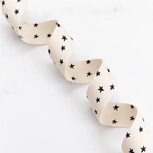 May include: Cream-coloured ribbon featuring a repeating pattern of black stars. The ribbon is curled, suggesting it's made of fabric. The white background provides a clean contrast. Suitable for crafts and gift wrapping. The ribbon's width is approximately 2.5 cm.