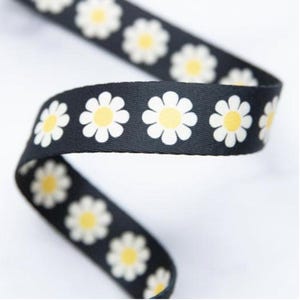 May include: Black ribbon featuring a repeating pattern of white daisies with yellow centres. The ribbon is curled, displaying the floral design. This decorative ribbon is suitable for crafts, gift wrapping, or adding a floral touch to various projects.