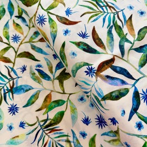 May include: White fabric with a watercolour botanical pattern. The design features green, blue, and brown leaves and small blue flower accents. The fabric is folded, showing the pattern's repeat.