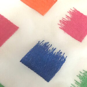 May include: A white fabric with embroidered squares in various colours, including orange, pink, blue, and green. The squares are filled with a textured, brushstroke-like pattern.