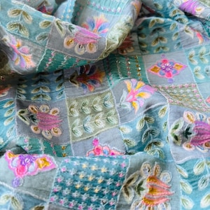 May include: Close-up of a fabric with a patchwork design. The fabric features a mix of squares in shades of blue, gray, and green. Each square is adorned with embroidered floral and leaf patterns in pink, yellow, and orange.