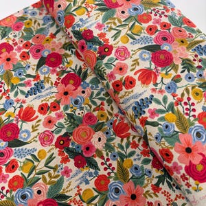May include: A cream-colored fabric with a floral pattern featuring red, pink, blue, and yellow flowers. The fabric is folded in half, showing the pattern on both sides.
