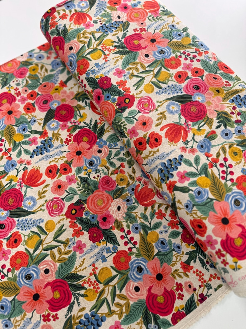 May include: A cream-colored fabric with a floral pattern featuring red, pink, blue, and yellow flowers. The fabric is folded in half, showing the pattern on both sides.