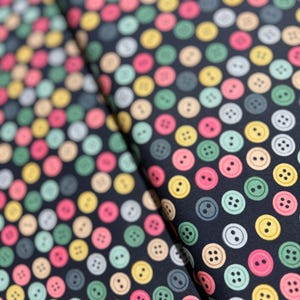 May include: A close-up of fabric featuring a repeating pattern of colourful buttons. The buttons are in shades of pink, yellow, green, grey, and white, set against a dark navy blue background. The fabric is folded, showing the pattern's continuity.
