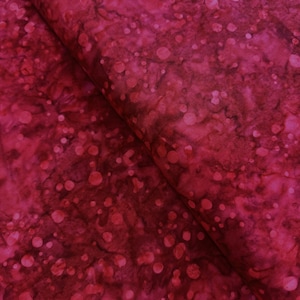 May include: Close-up of a folded fabric with a deep red, mottled pattern. The fabric features a speckled design with varying sizes of circular shapes, creating a textured appearance. The overall effect is a rich, burgundy color.